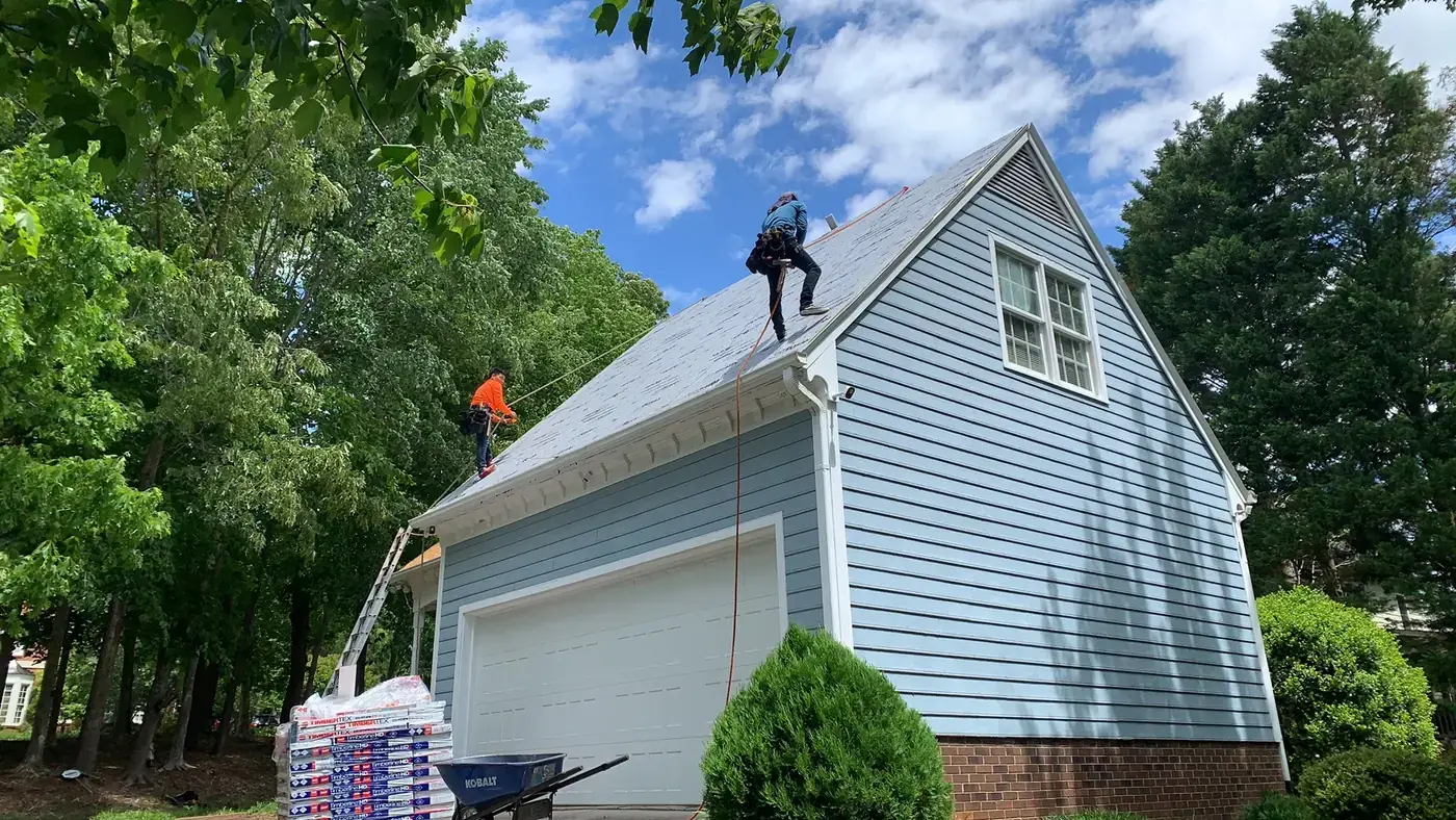 Two roofers roped off on a steep slope laying shingles, with bundles stacked in the driveway below